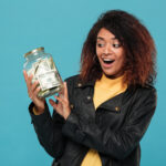 Surprised happy african woman in leather jacket looking at jar with money over blue background